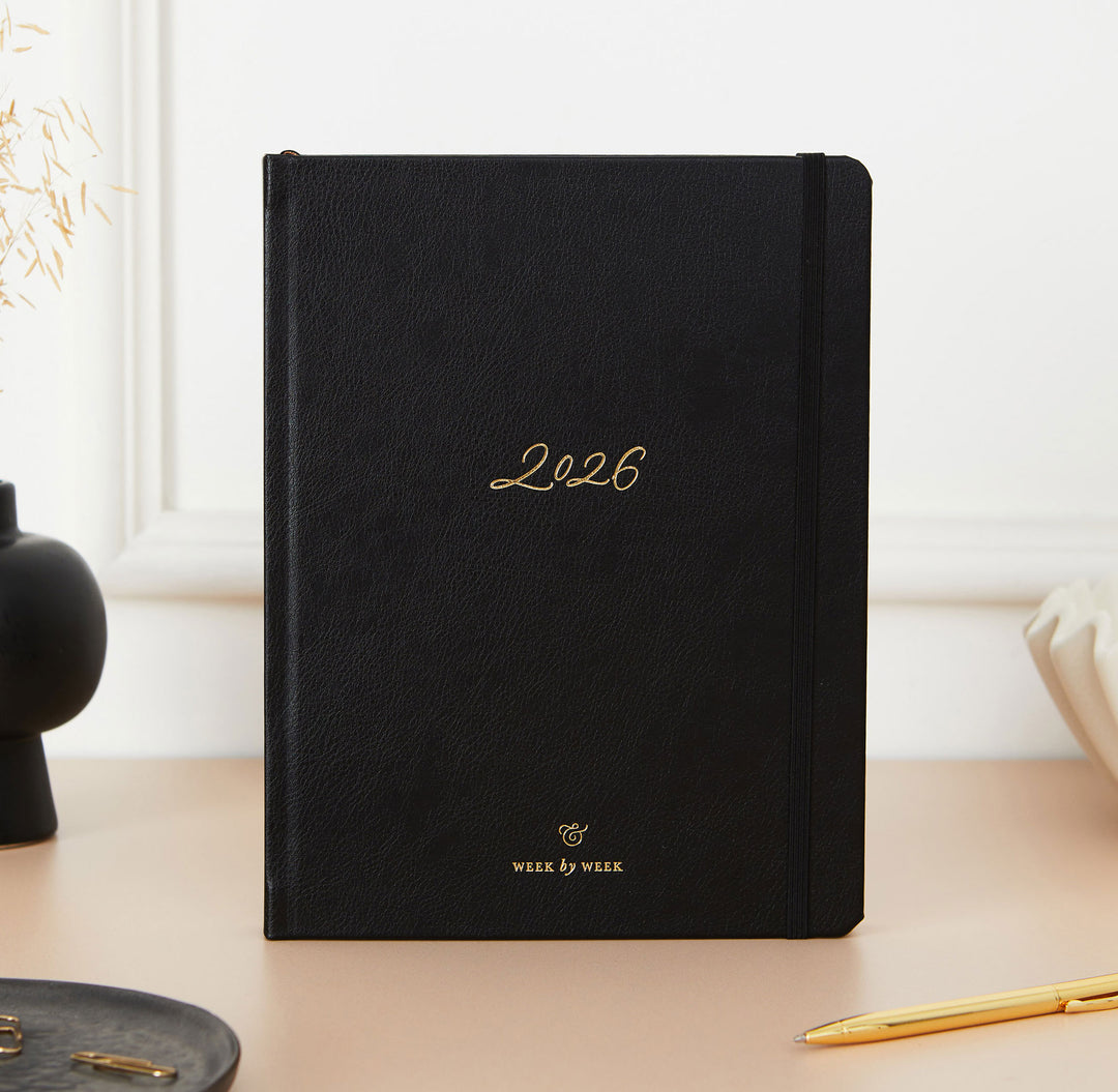 Black planner with '2026' embossed on it, placed on a desk with a pen and decorative items.