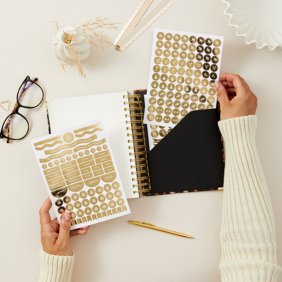 Person holding gold planner stickers next to a planner on a light surface.