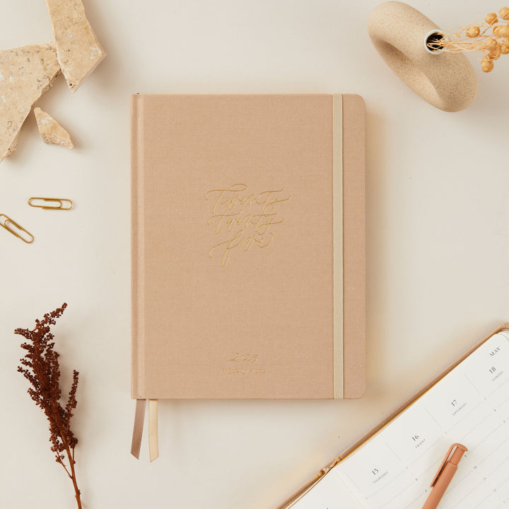 2025 planner with an ivory fabric cover and elegant gold details, featuring weekly time-blocking with equal space for each day, to-do lists, and monthly budget and goal sections.