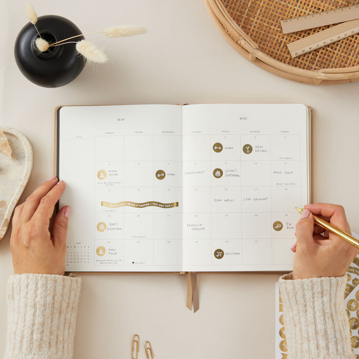 Ivory fabric-covered 2025 planner with gold accents, offering time-blocking space for each day of the week, to-do lists, notes, and sections for monthly goals and budgeting.