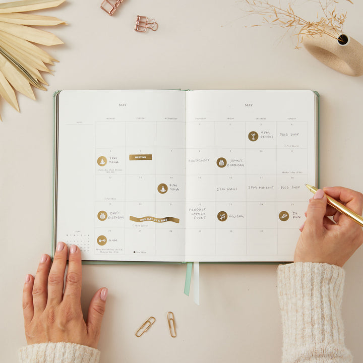 Stylish 2025 weekly planner with a sage green vegan leather cover and gold details, featuring time-blocking for each day of the week, to-do lists, and monthly goals and budgeting pages.
