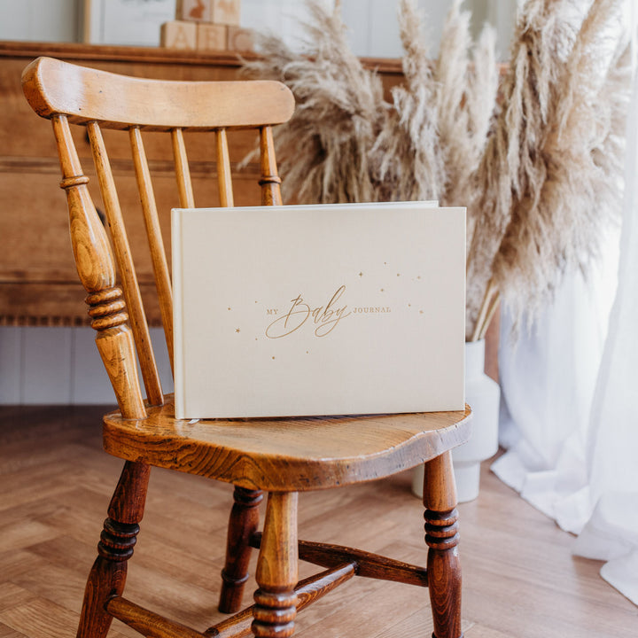 Cover of a baby memory book with an elegant ivory background and gold accents, featuring delicate illustrations of stars, hearts, and a central title that reads 'My Baby Journal' in stylish gold lettering