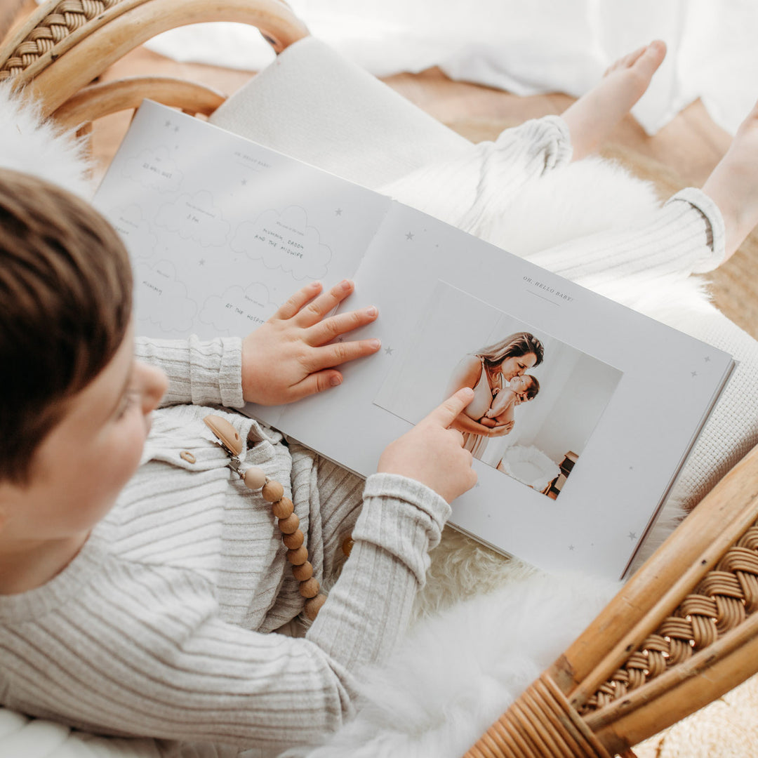 Young child sitting on a cozy chair, looking through a baby memory book with an ivory cover and gold accents, pages filled with photos.