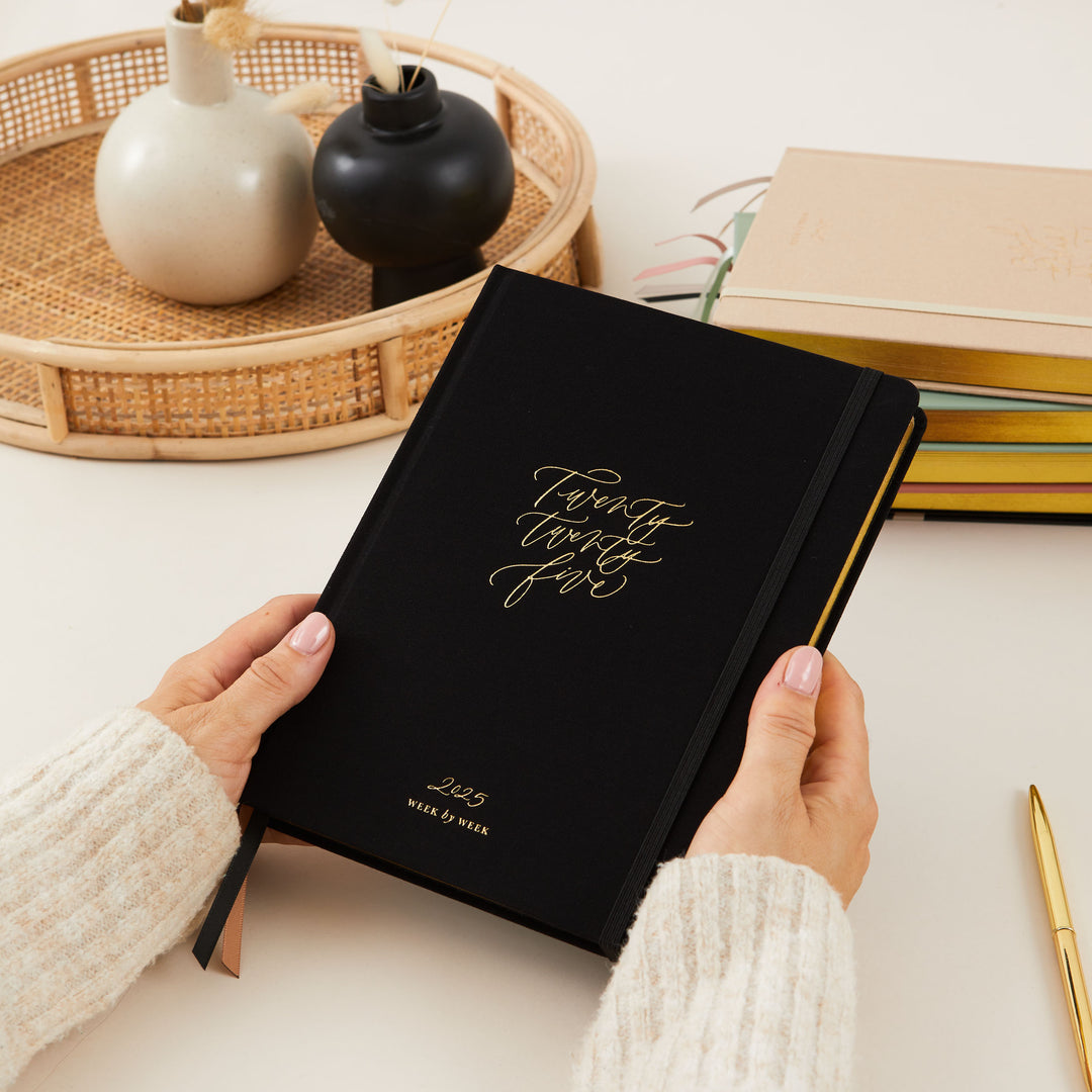 Stylish 2025 weekly planner with a soft black vegan leather cover and gold accents, featuring time-blocking for each day of the week, to-do lists, and sections for notes and budgeting.
