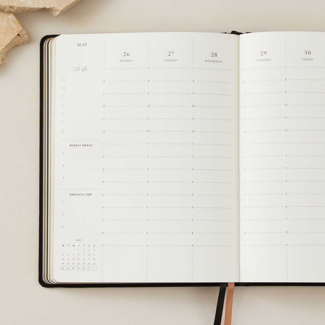 Stylish 2025 weekly planner with a soft black vegan leather cover and gold accents, featuring time-blocking for each day of the week, to-do lists, and sections for notes and budgeting.
