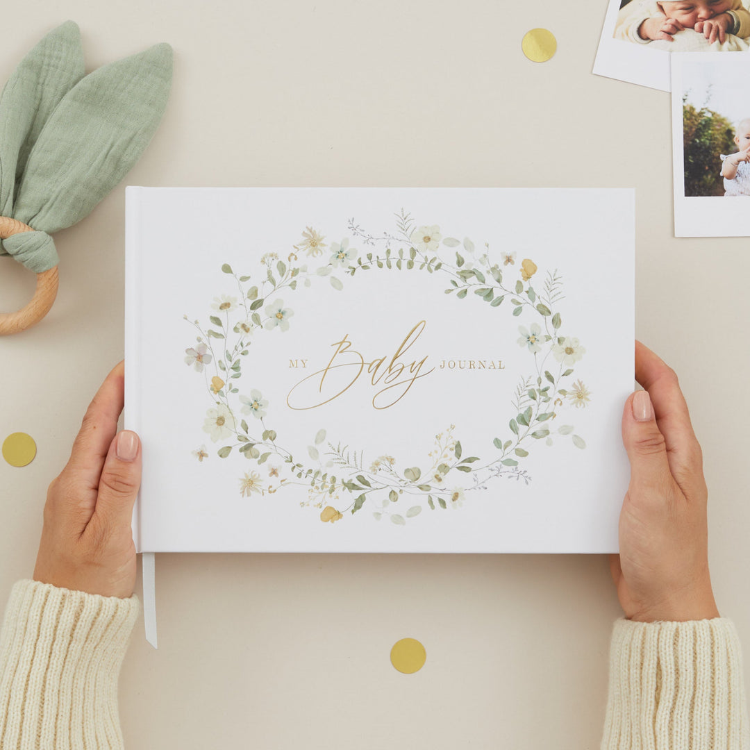 Person holding a baby album with floral design and 'It's a Baby' text, surrounded by baby items on a light background.