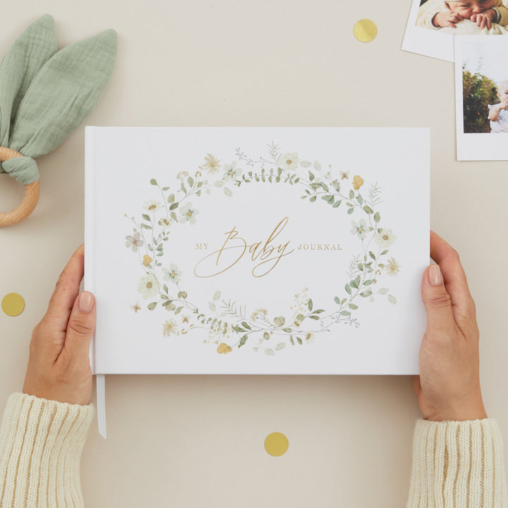 Person holding a baby album with floral design and 'It's a Baby' text, surrounded by baby items on a light background.