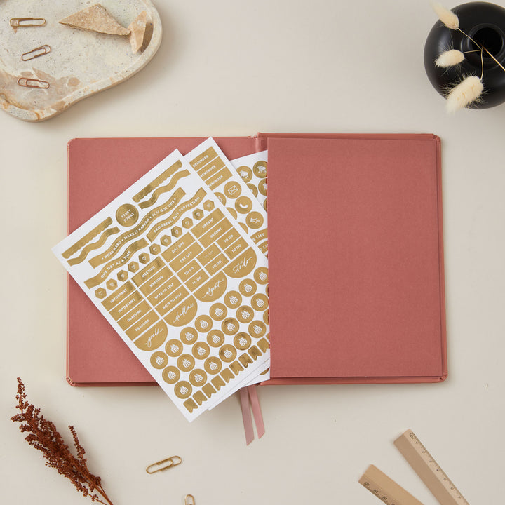 Elegant 2025 planner with a clay vegan leather cover and gold details, featuring time-blocking for each day, to-do lists, and sections for monthly goals and budget tracking.