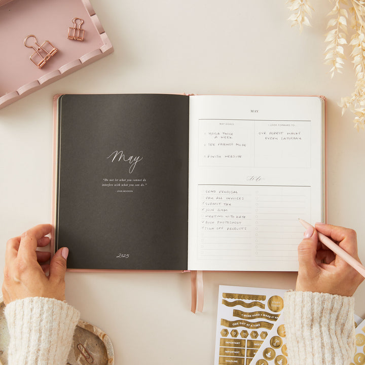 Stylish 2025 weekly planner with a soft pink vegan leather cover and gold accents, featuring time-blocking for each day of the week, to-do lists, and sections for notes and budgeting.