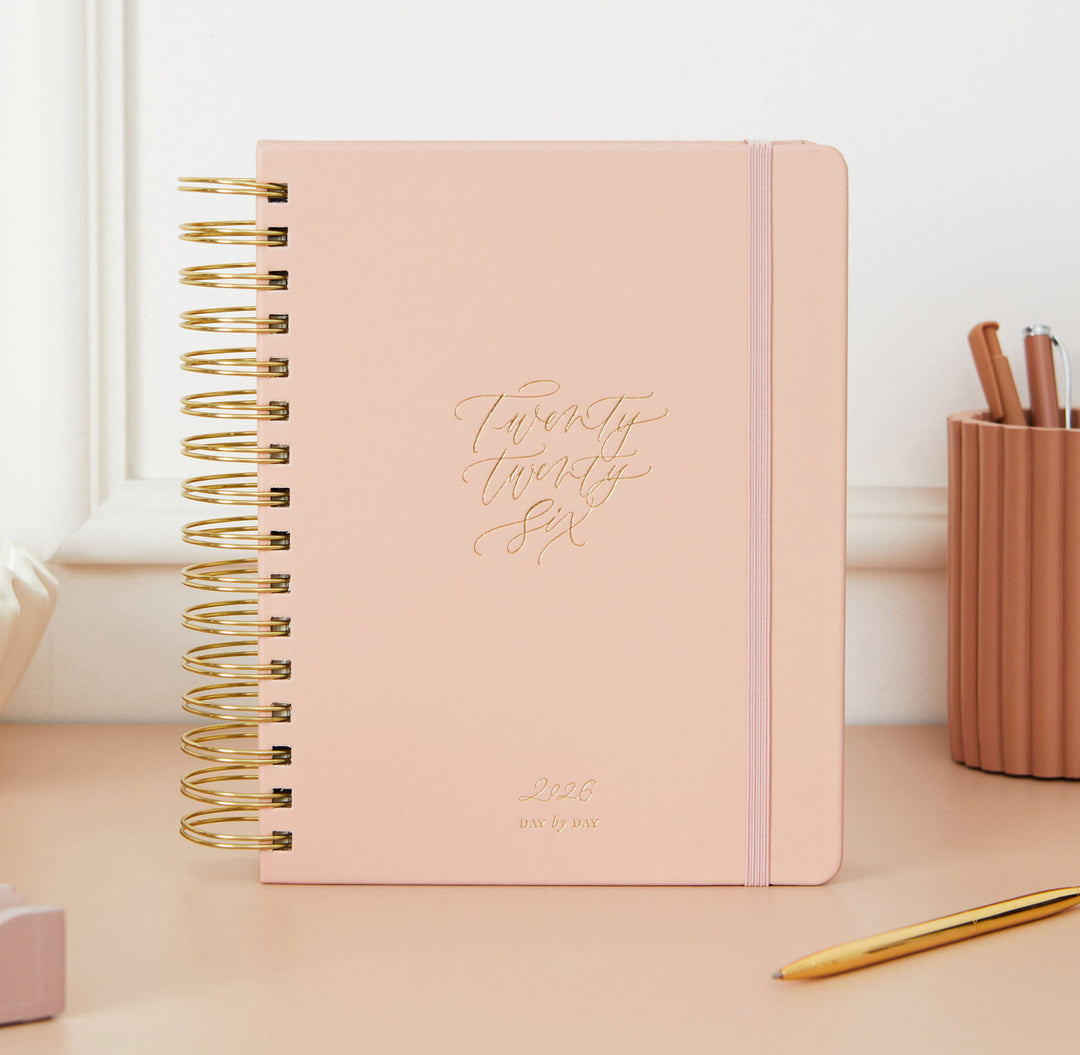 Pink planner with gold text on a desk with a pen holder and pen.