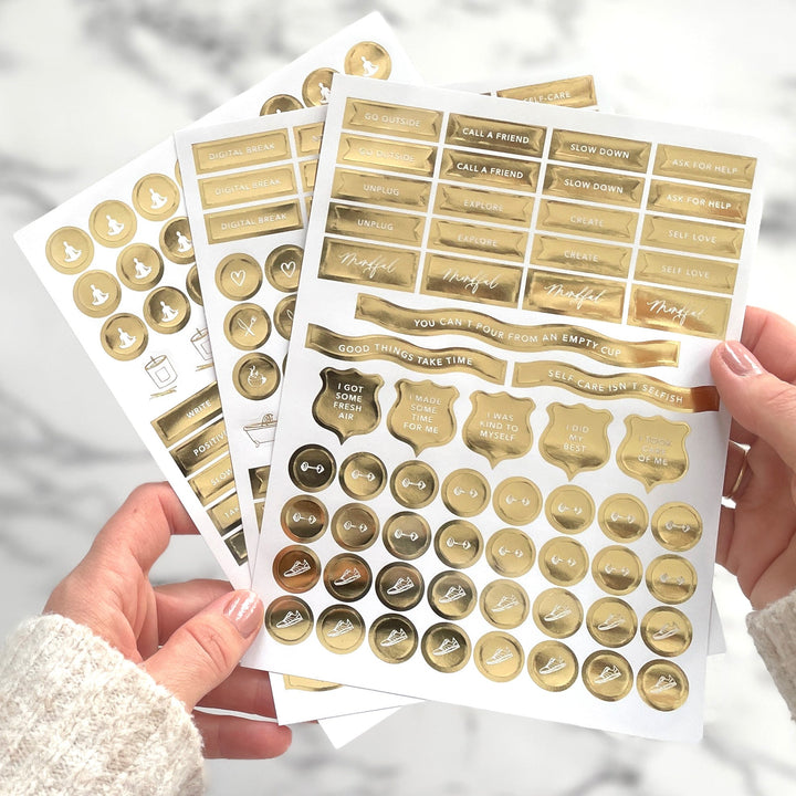 Hands holding three gold foil motivational sticker sheets with phrases like "I took care of me" and "Self-care isn't selfish"