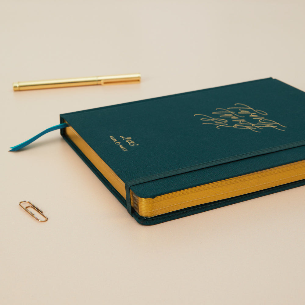 Green notebook with gold accents on a beige surface, accompanied by a pen and paperclip.