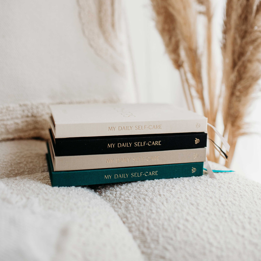 Four My Daily Self-Care journals stacked in cream, black, taupe and green linen with gold foil spines, on a cosy boucle chair