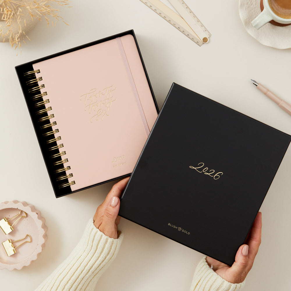 Person holding a gift box with a '2026' pink planner inside, on a light surface.