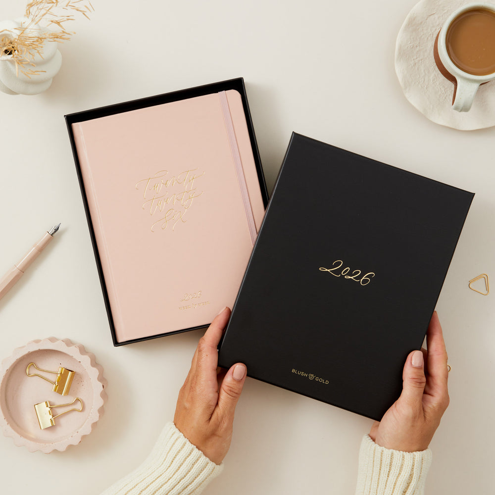 Person holding a black planner with '2026' on a light surface with a cup of coffee and decorative items.