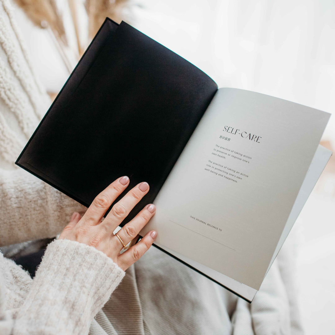 Hand resting on an open journal showing a self-care definition and a "this journal belongs to" line for a personal touch