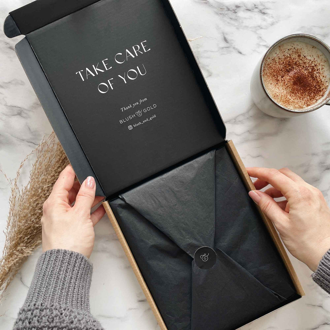 Hands opening a Blush & Gold box printed with "Take care of you", with black tissue sealed by a gold ampersand sticker and a cappuccino on marble