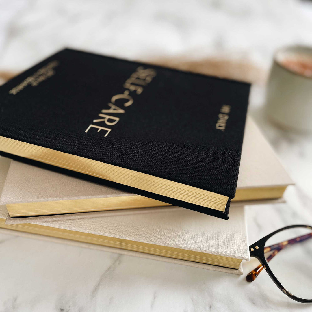 Black linen self-care journal with gold gilded page edges resting on two cream journals, next to tortoiseshell glasses