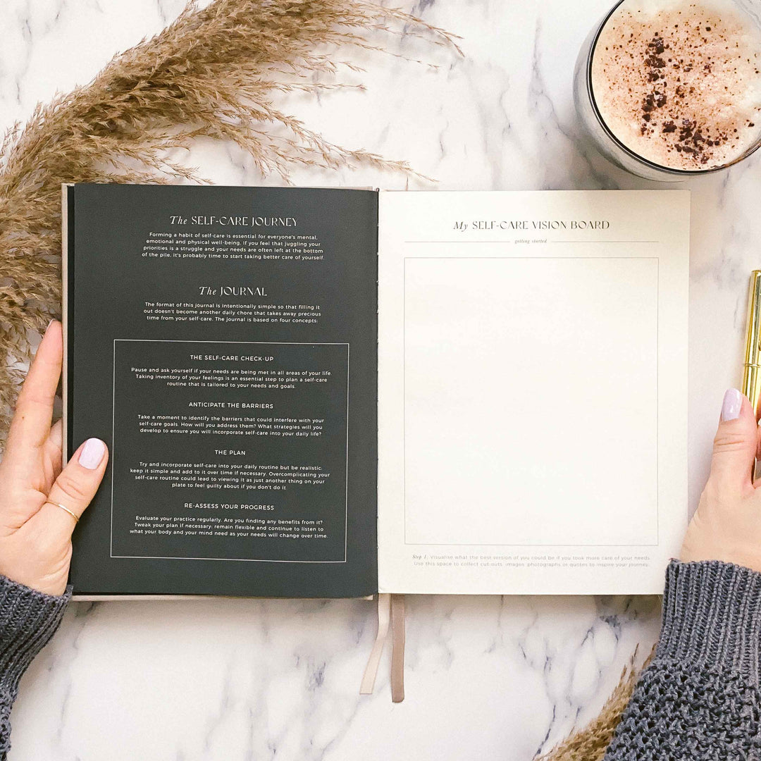 Journal open to the self-care journey guide on a dark page and a blank vision board page, beside pampas grass and a hot chocolate