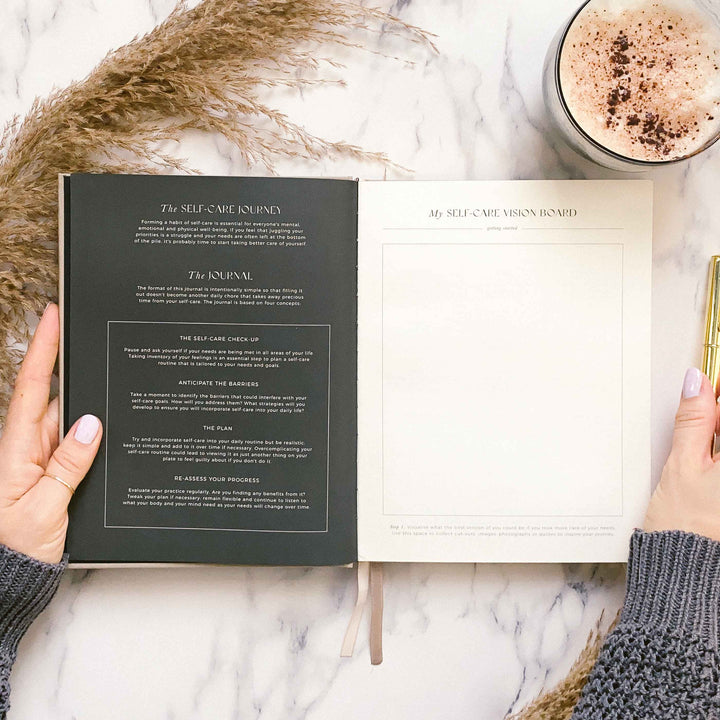 Journal open to the self-care journey guide on a dark page and a blank vision board page, beside pampas grass and a hot chocolate
