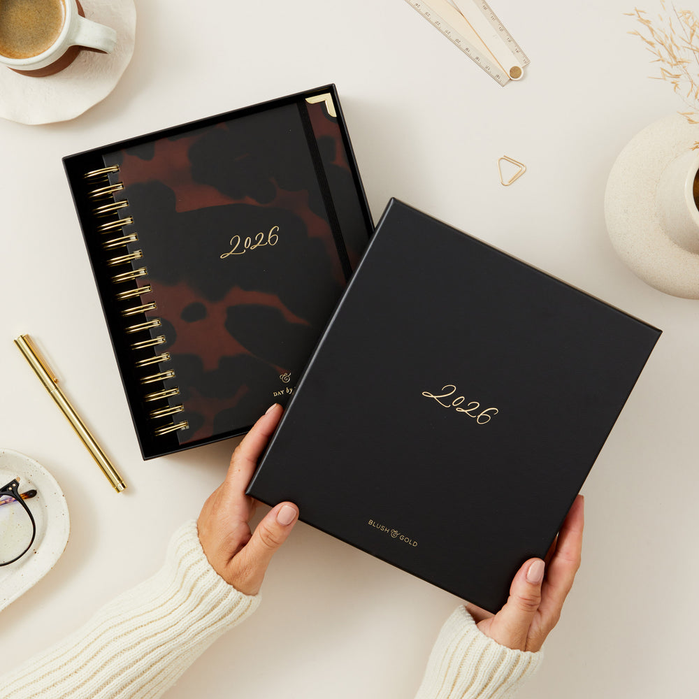 Person holding a planner with '2026' on a desk with a cup of coffee and stationery.