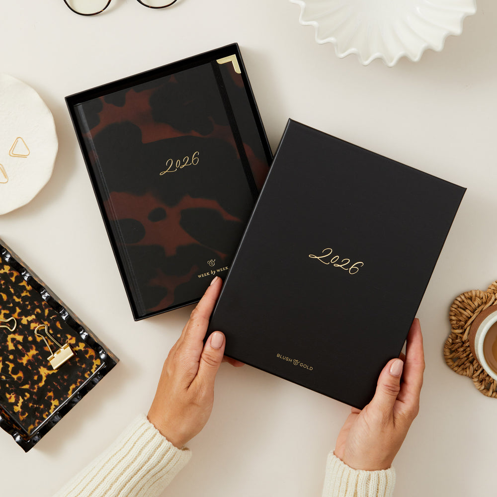 Person holding a black planner with '2026' on a light surface with decorative items.