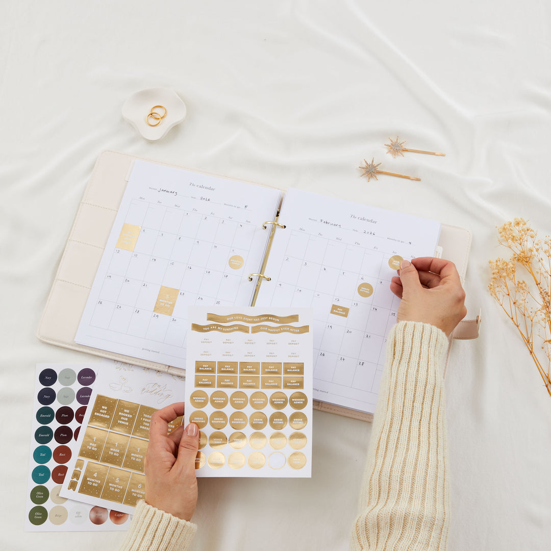 Open wedding planner binder showing monthly calendar pages with gold foil sticker sheets being held up in front