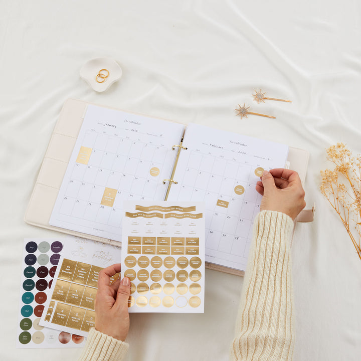 Open wedding planner binder showing monthly calendar pages with gold foil sticker sheets being held up in front