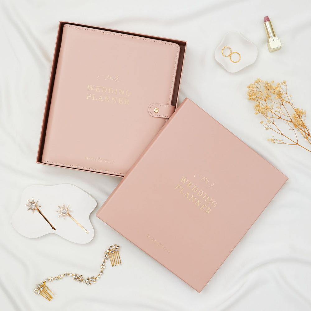 Blush pink wedding planner binder inside an open gift box alongside a matching box lid and bridal accessories flatlay including hair comb and star hair pins