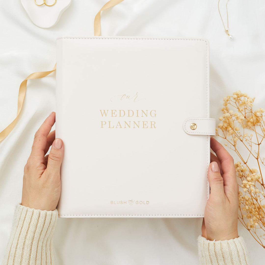 Hands holding an ivory leather wedding planner binder with gold foil lettering, styled with wedding rings and dried flowers