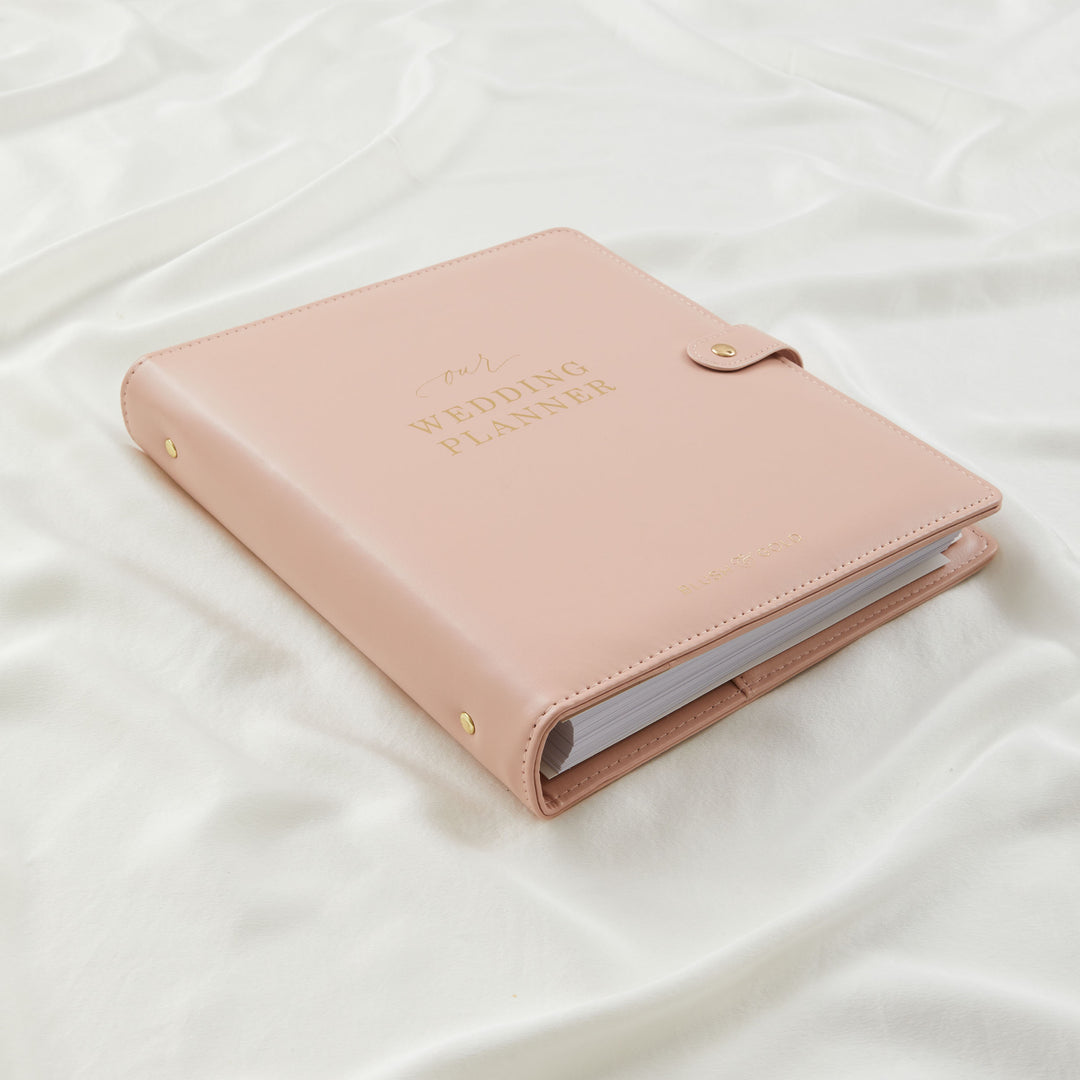 Blush pink vegan leather wedding planner binder closed on white silk with gold foil cover text and gold stud details