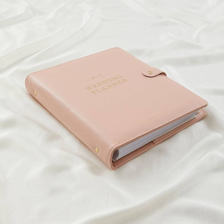 Blush pink vegan leather wedding planner binder closed on white silk with gold foil cover text and gold stud details