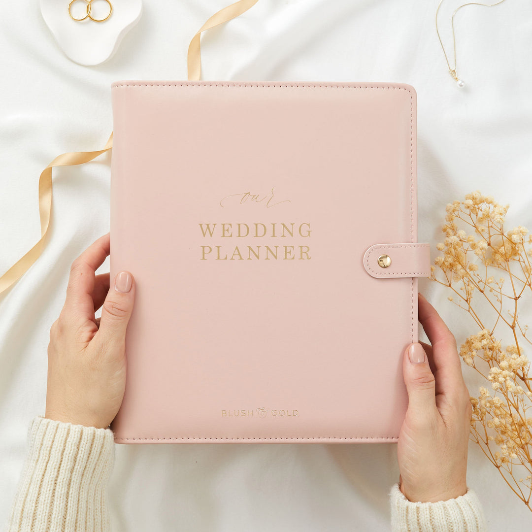 Two hands holding a blush pink wedding planner binder from below in an overhead flatlay shot with gold ribbon and dried flowers