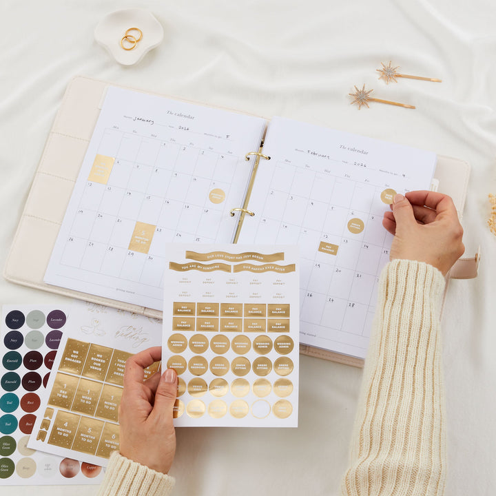 Open wedding planner binder showing monthly calendar pages with gold foil milestone sticker sheets and colour dot sticker sheet spread out below