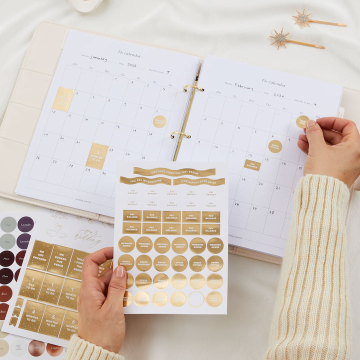 Hands placing gold foil stickers onto wedding planner calendar pages showing January and February with milestone and task sticker sheets spread out below
