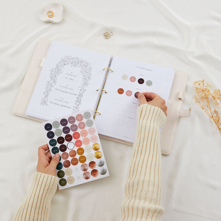 Hands placing colour dot stickers onto a wedding planner colour scheme page with a full sticker sheet of named colour options held below