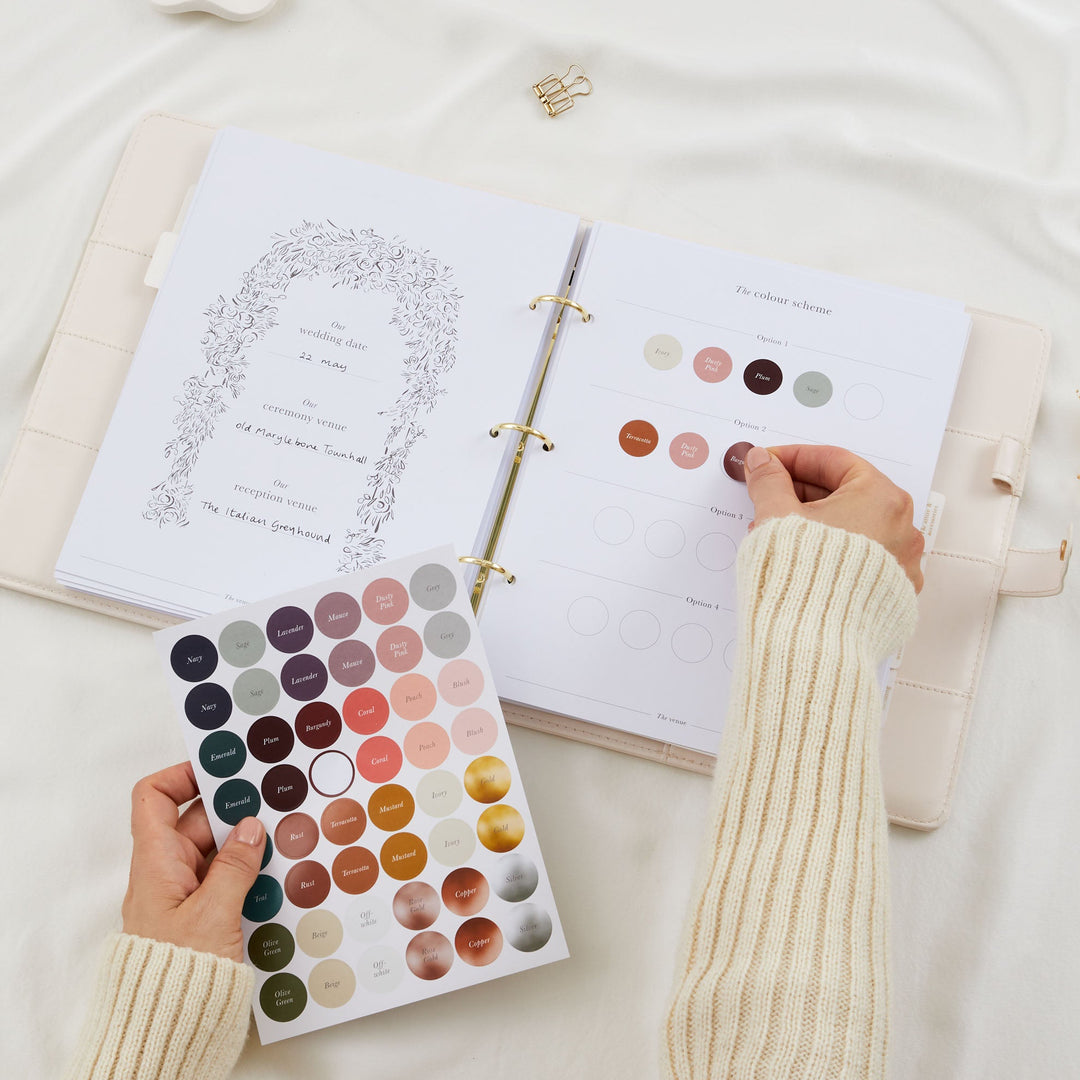 Hands placing a colour dot sticker onto a wedding planner colour scheme page with a full named colour sticker sheet held below and completed wedding date page visible