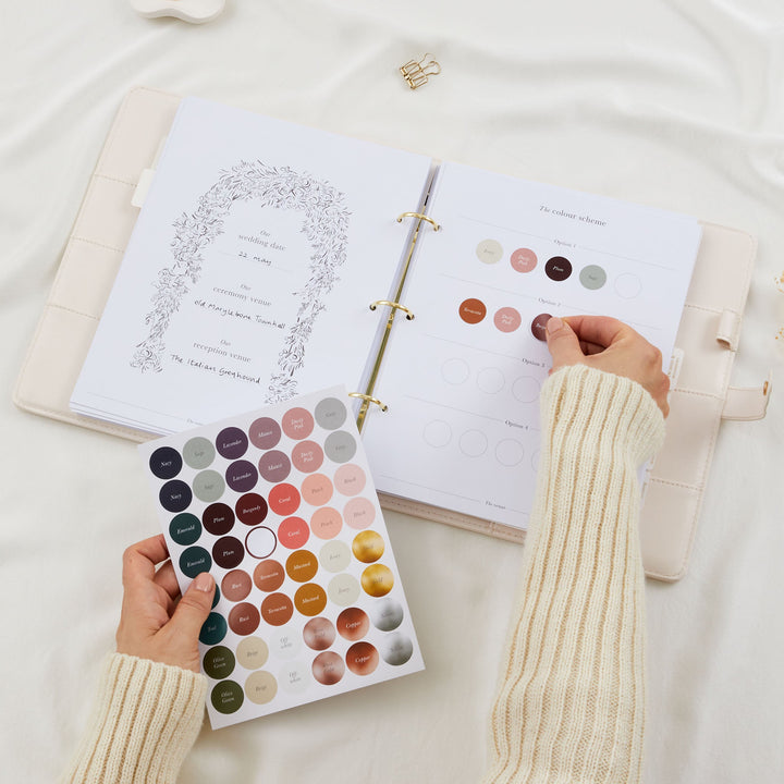 Hands placing colour dot stickers onto a wedding planner colour scheme page with a full named colour sticker sheet held below and wedding date page visible