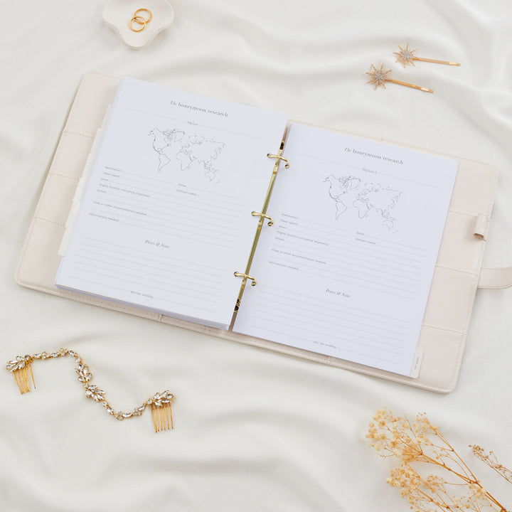 Open wedding planner binder showing two honeymoon research comparison pages with world map illustrations and fields for destination, travel agent and flight details