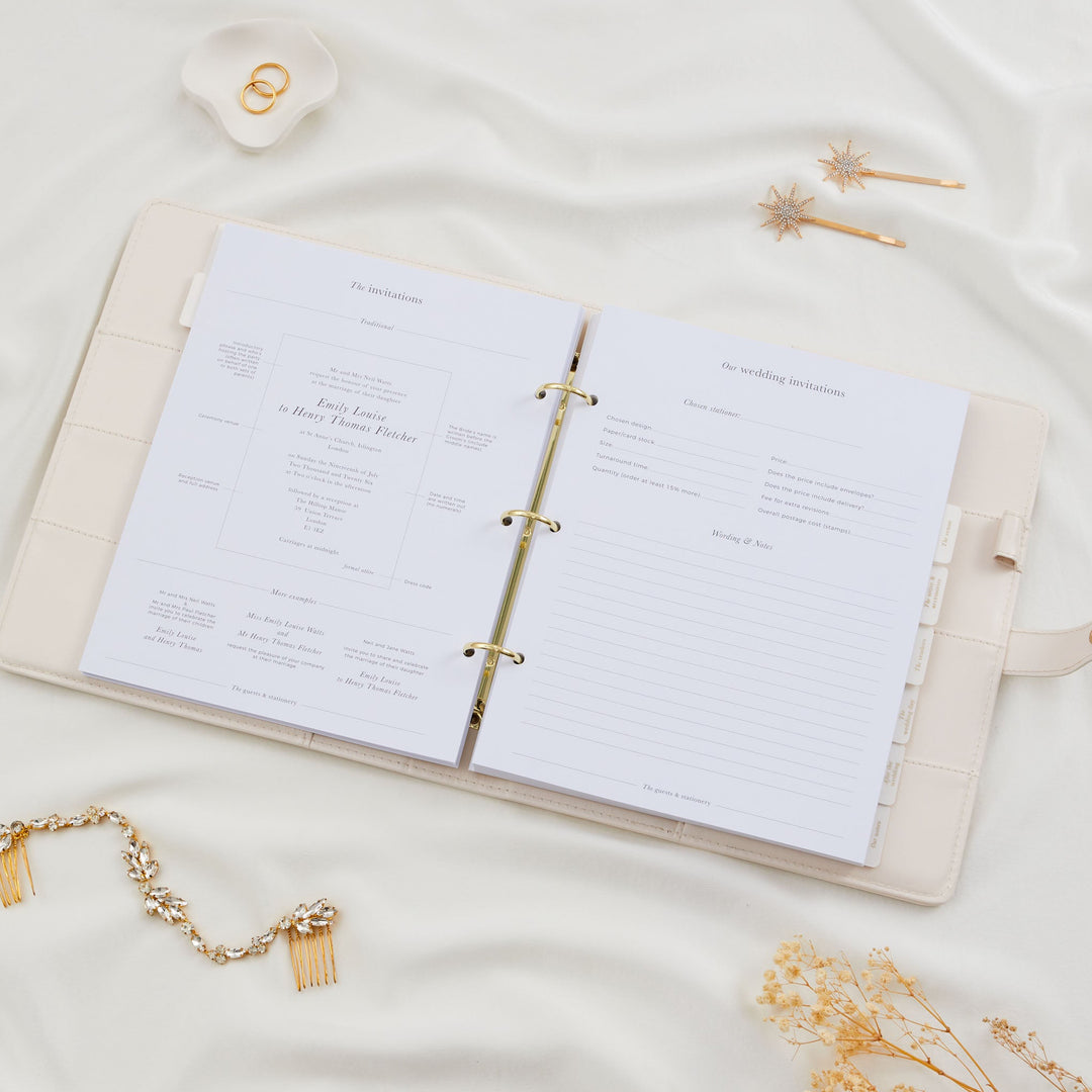 Open wedding planner binder showing a wedding invitations section with traditional wording examples and a stationer comparison form styled with a bridal hair comb flatlay