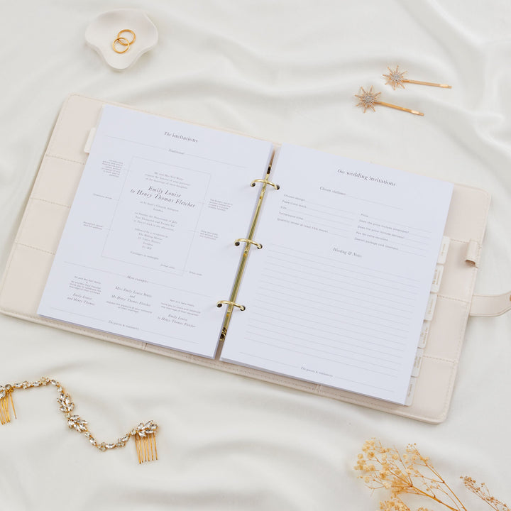 Open wedding planner binder showing a wedding invitations section with traditional wording examples and a stationer comparison form styled with a bridal hair comb flatlay
