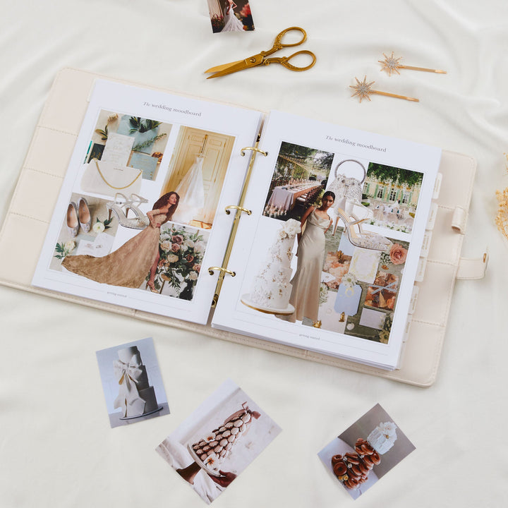 Open wedding planner binder showing filled wedding moodboard pages with bridal inspiration photos alongside gold scissors and loose cake photos scattered below