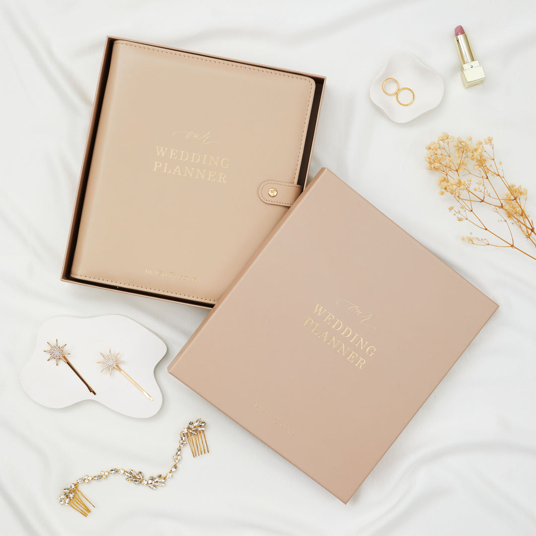 Nude leather wedding planner binder in open gift box styled with gold bridal hair accessories dried flowers and rings on white linen