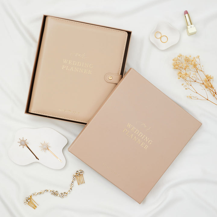 Nude leather wedding planner binder in open gift box styled with gold bridal hair accessories dried flowers and rings on white linen