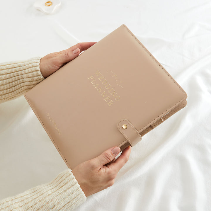 Hands holding a nude leather wedding planner binder with gold foil lettering and snap closure