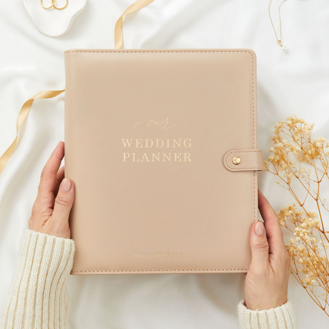 Hands holding a nude tan leather wedding planner binder styled with gold satin ribbon wedding rings and dried flowers on white linen