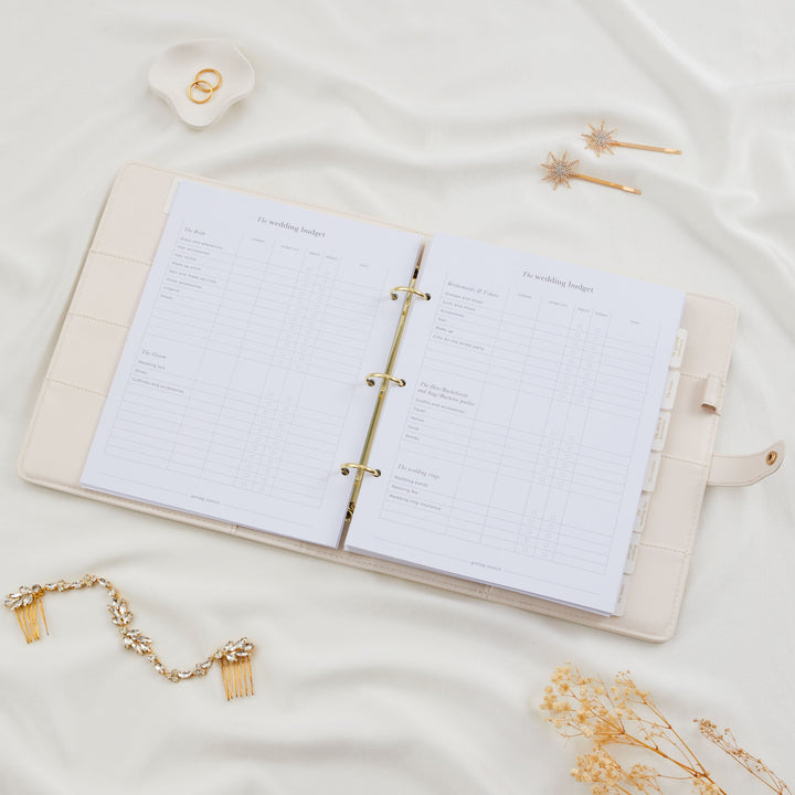 Open ivory wedding planner binder showing wedding budget worksheet pages with gold ring binder hardware, styled with bridal hair combs and dried flowers