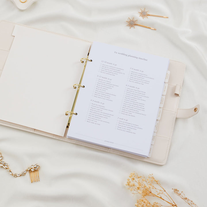 Open ivory wedding planner binder displaying a wedding planning timeline checklist, styled with bridal accessories and dried flowers