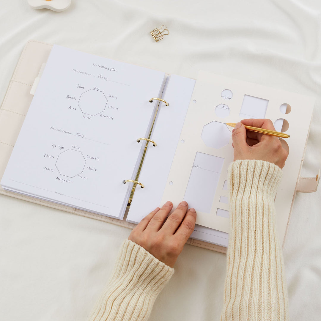 Close up of hands using a table stencil and gold pen to draw a seating plan in a wedding planner binder with completed named table diagrams visible