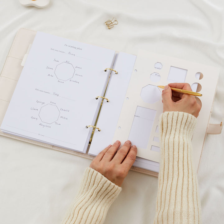 Close up of hands using a table stencil and gold pen to draw a seating plan in a wedding planner binder with completed named table diagrams visible